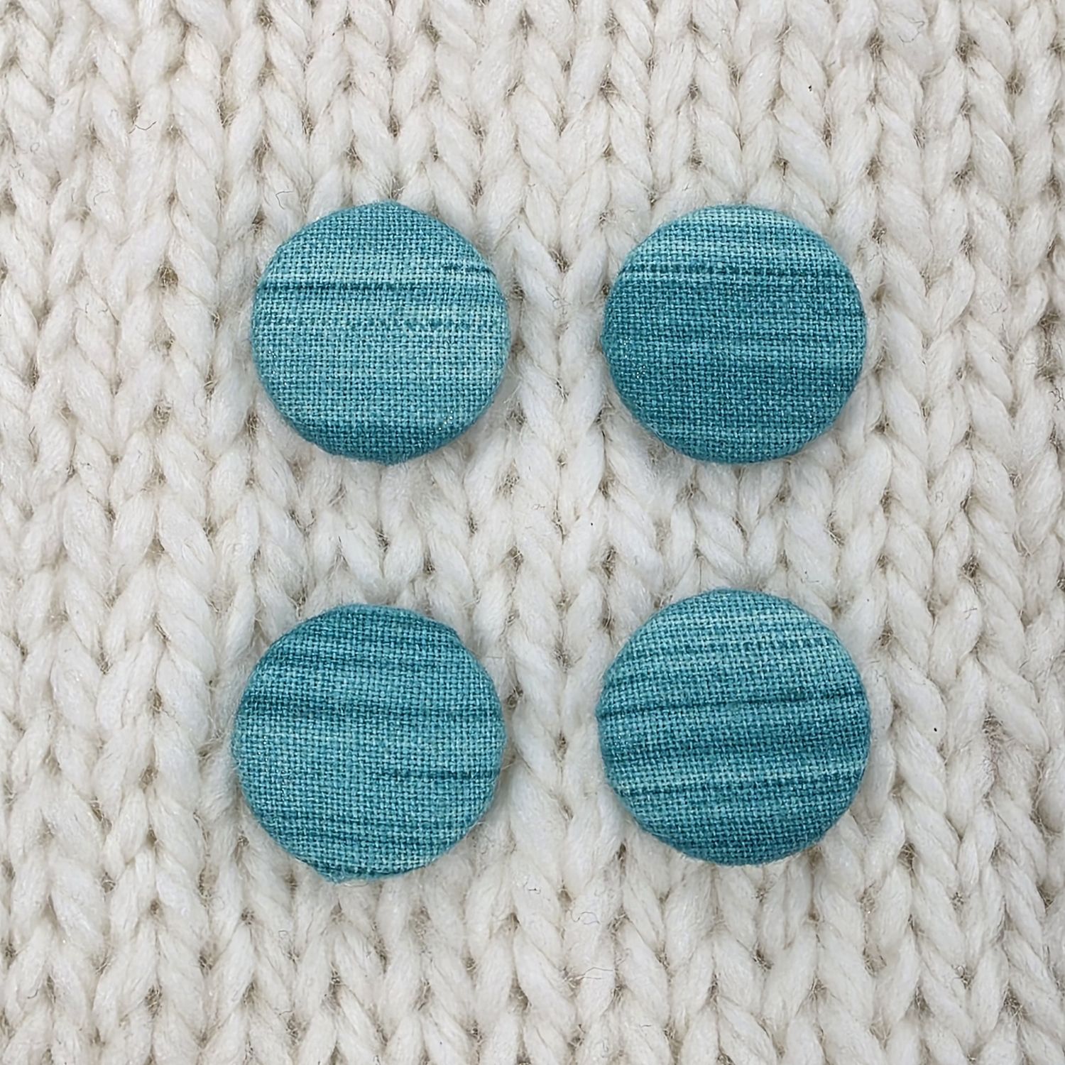 4 teal fabric covered buttons with a slight striped print, on a cream knitted background.