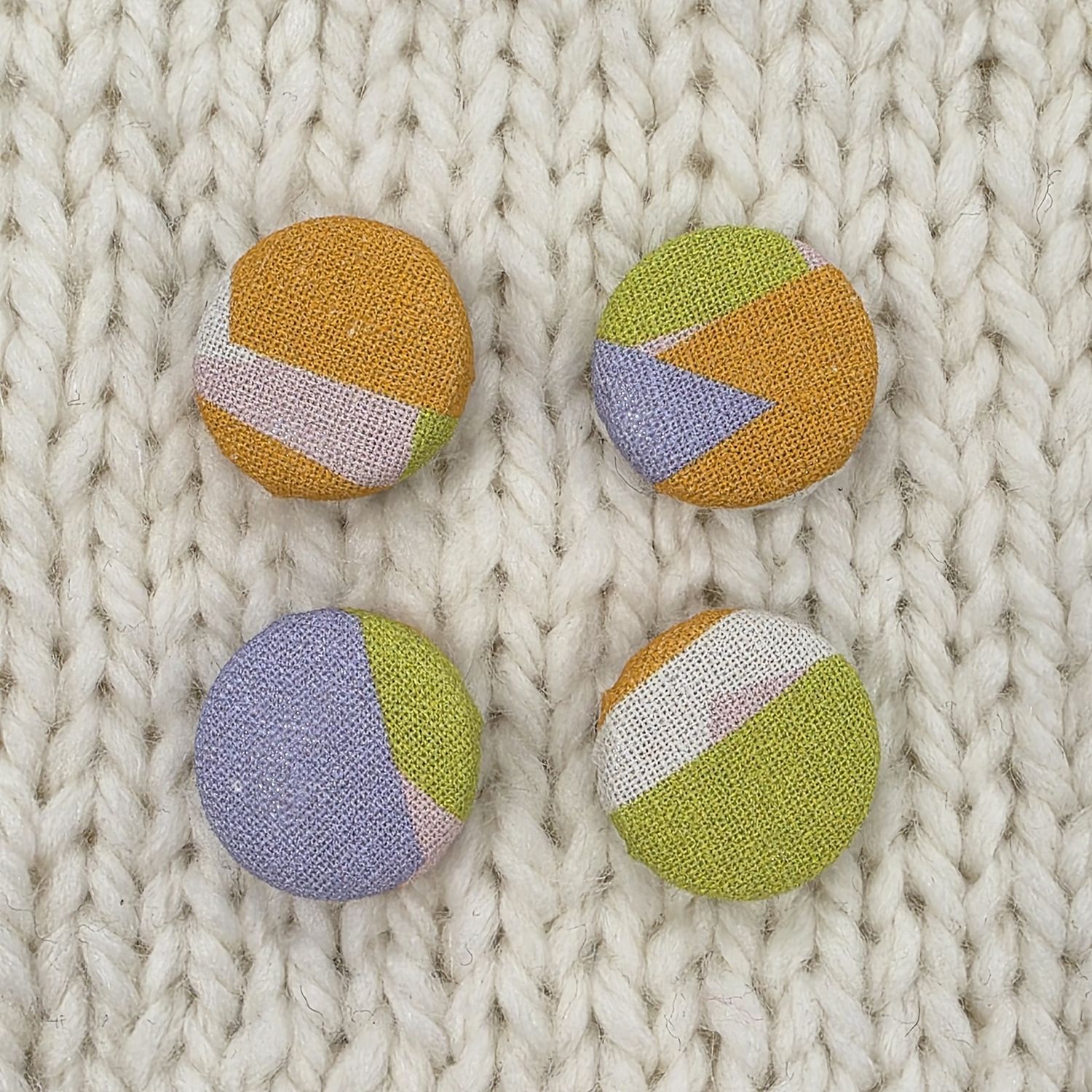 4 white fabric covered buttons with orange, purple and green geometric print, on a cream knitted background.