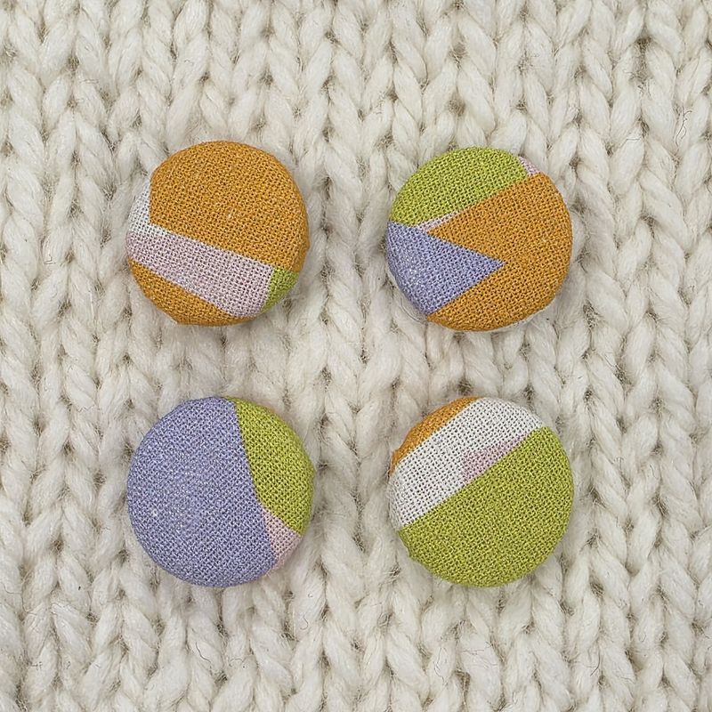 4 white fabric covered buttons with orange, purple and green geometric print, on a cream knitted background.