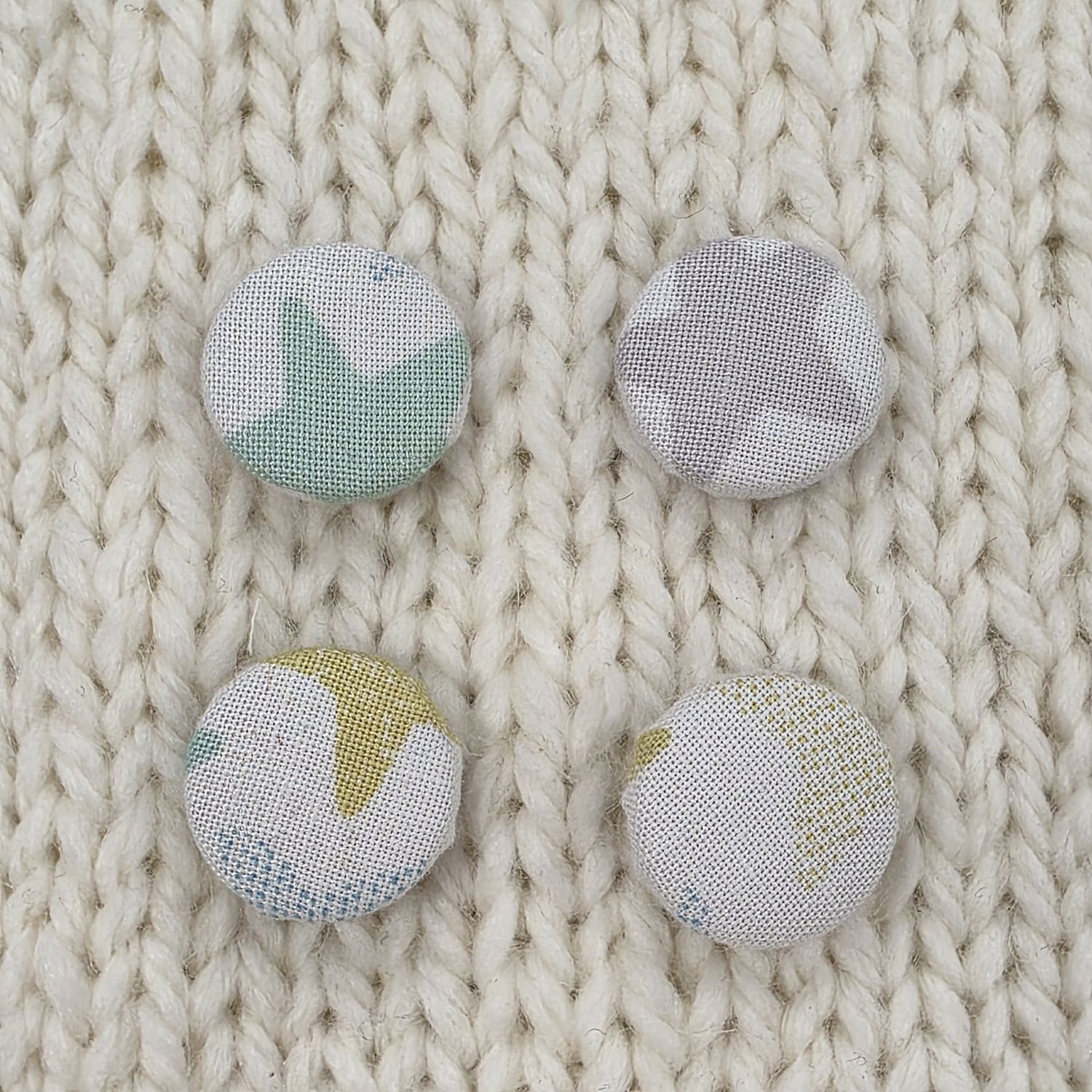 4 white fabric covered buttons with grey, green and mint blue star print, on a cream knitted background.