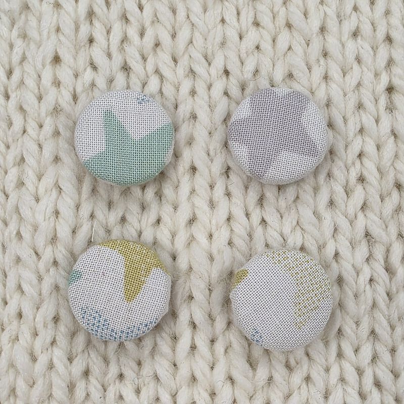 4 white fabric covered buttons with grey, green and mint blue star print, on a cream knitted background.