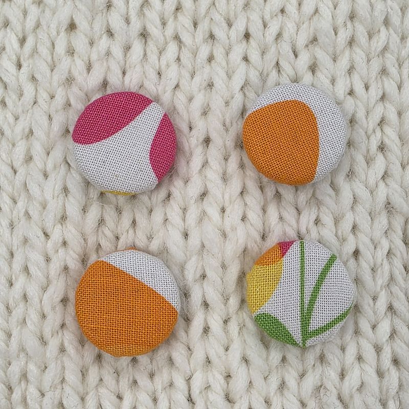4 white fabric covered buttons with pink, orange, yellow and green abstract print, on a cream knitted background.