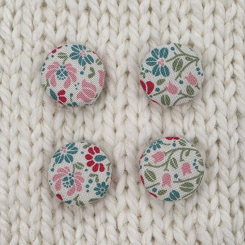 4 white fabric covered buttons with pink, green and blue floral print, on a cream knitted background.