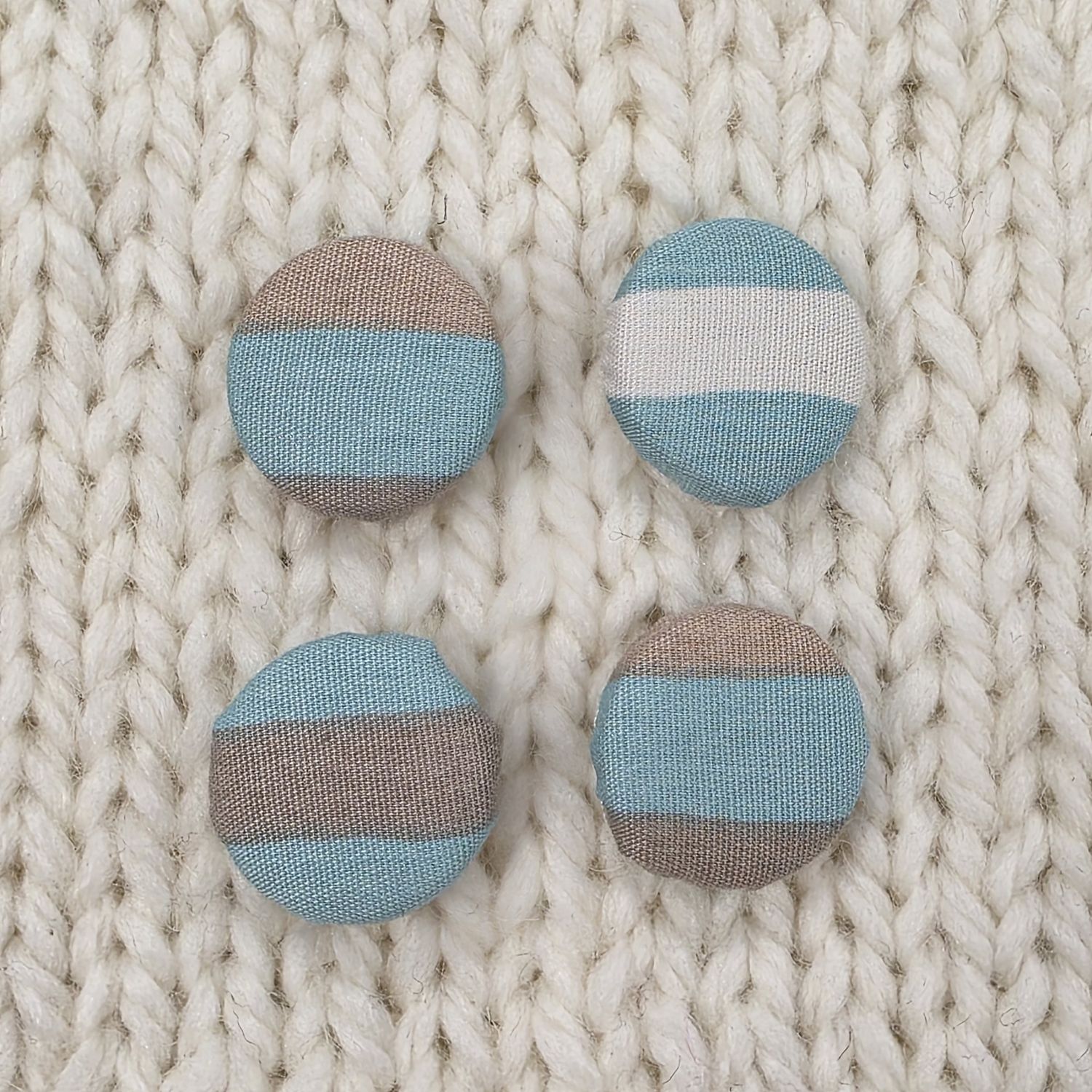 4 aqua blue fabric covered buttons with wide white and grey stripes, on a cream knitted background.
