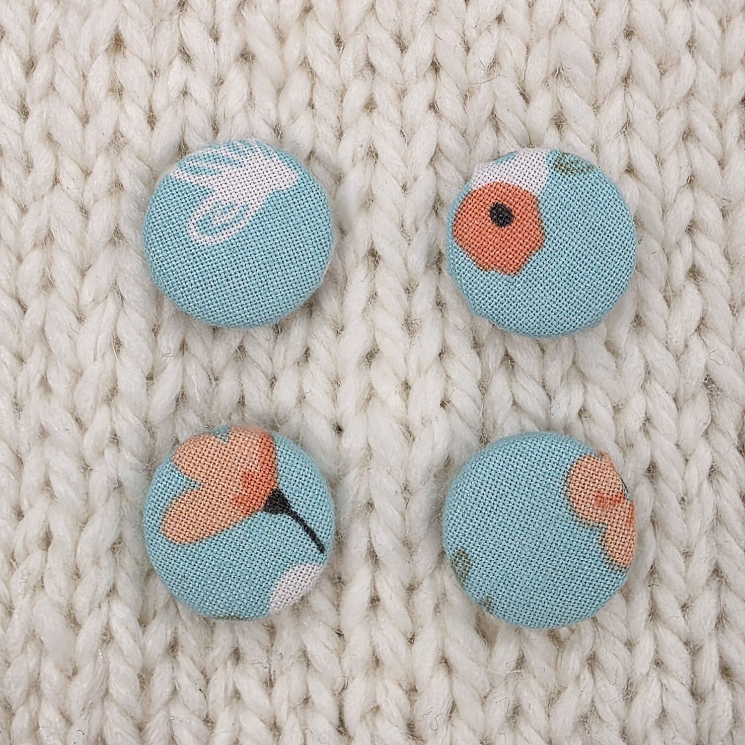 4 light blue fabric covered buttons with white and pink floral print, on a cream knitted background.