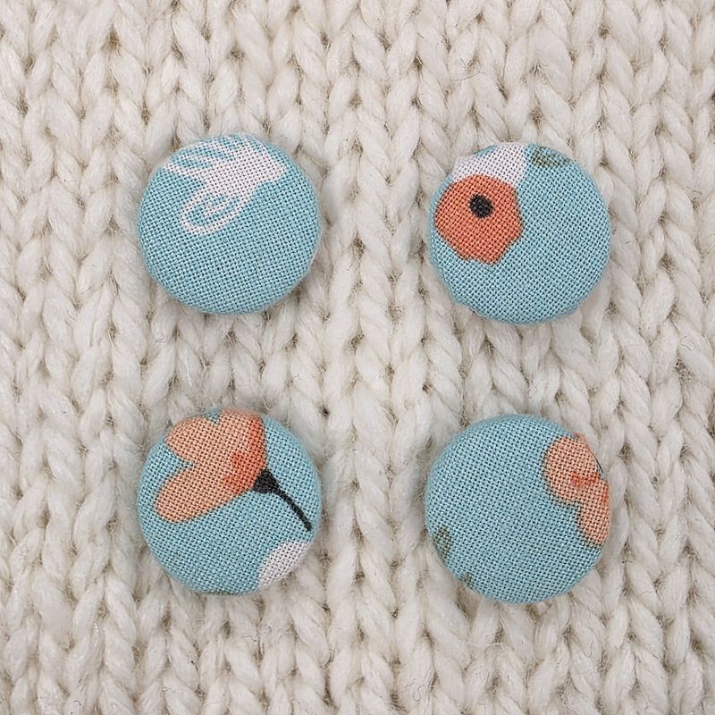 4 light blue fabric covered buttons with white and pink floral print, on a cream knitted background.