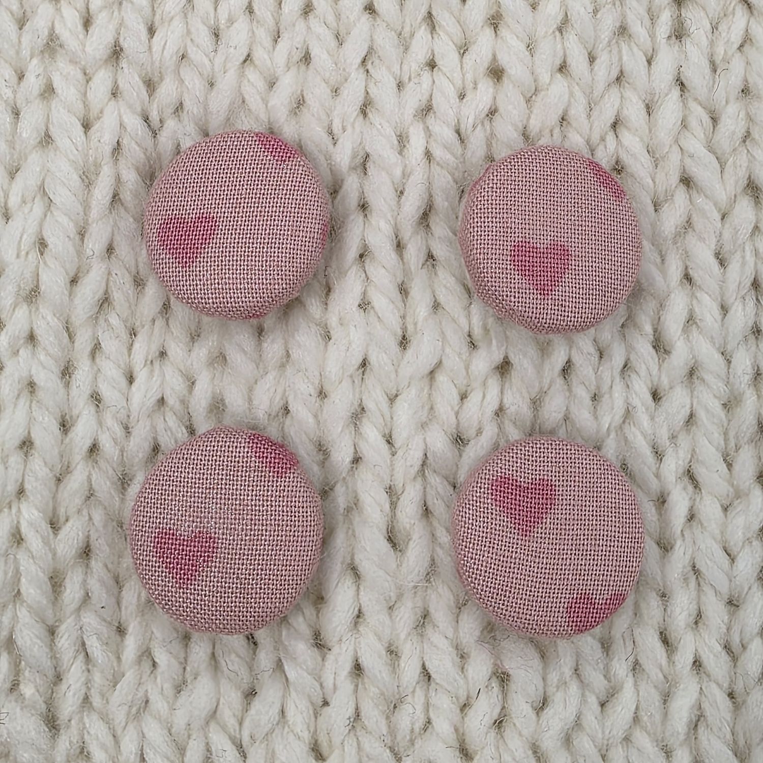 4 light pink fabric covered buttons with dark pink heart print, on a cream knitted background.