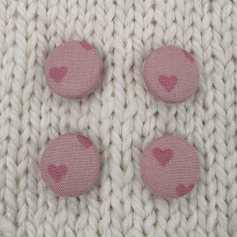 4 light pink fabric covered buttons with dark pink heart print, on a cream knitted background.