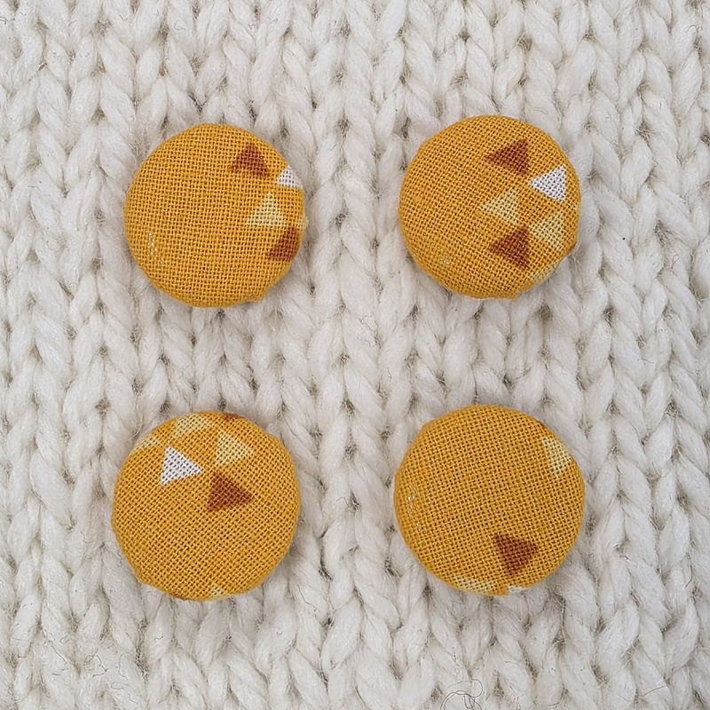 4 orange fabric covered buttons with dark orange and white triangle print, on a cream knitted background.