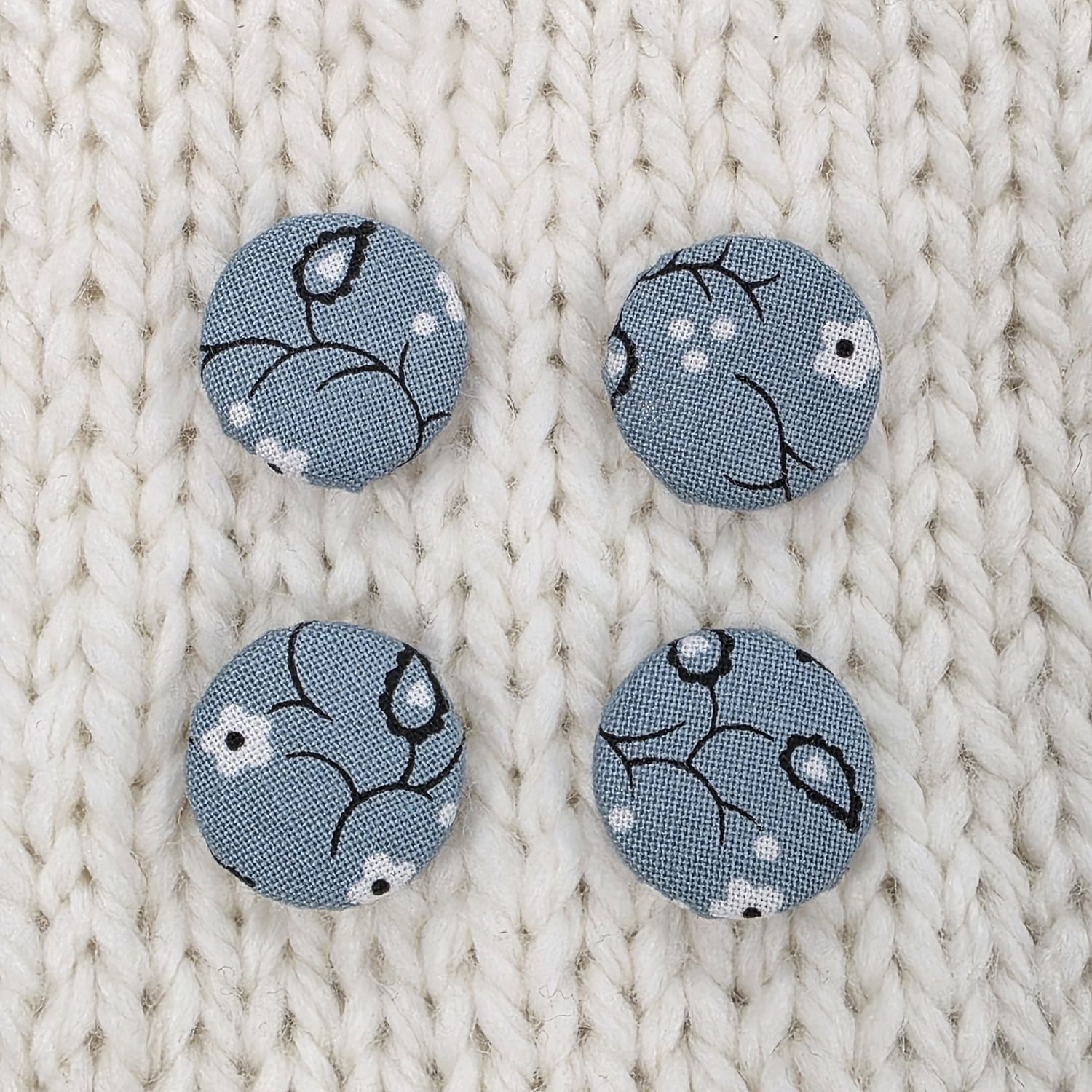 4 dusky blue fabric covered buttons with white and dark grey floral print, on a cream knitted background.
