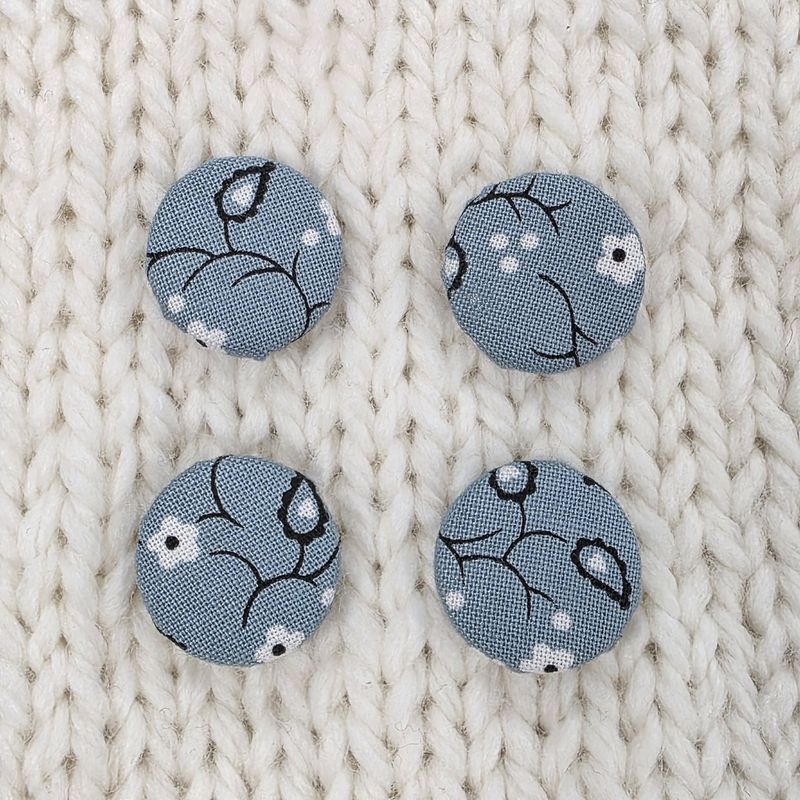 4 dusky blue fabric covered buttons with white and dark grey floral print, on a cream knitted background.