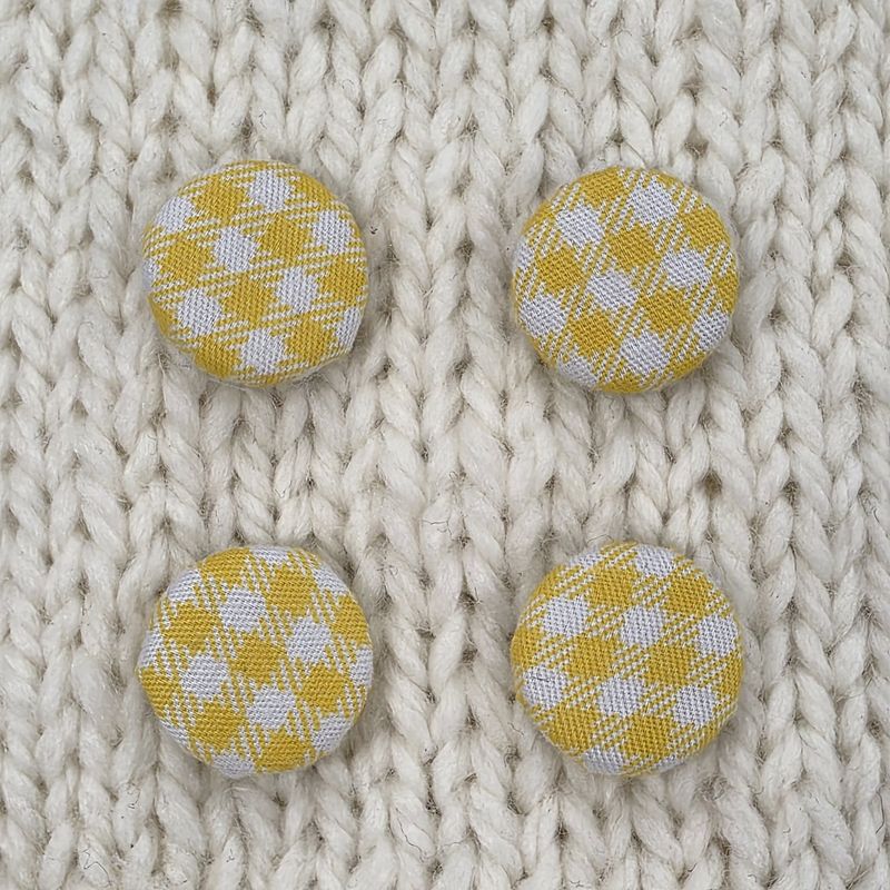 4 fabric covered buttons with a yellow and white checked print, on a cream knitted background.