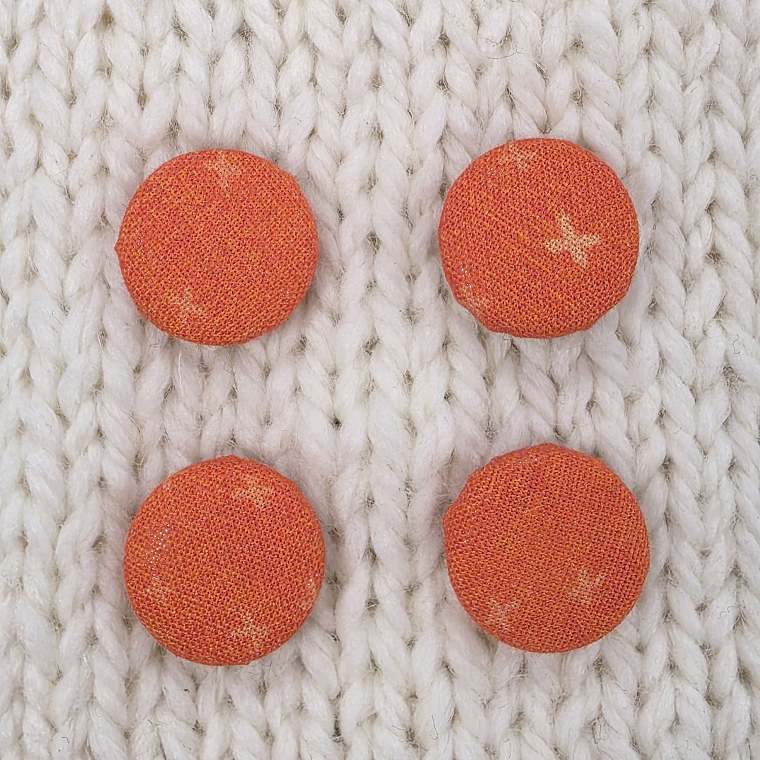 4 dark orange fabric covered buttons with light orange crosses print, on a cream knitted background.