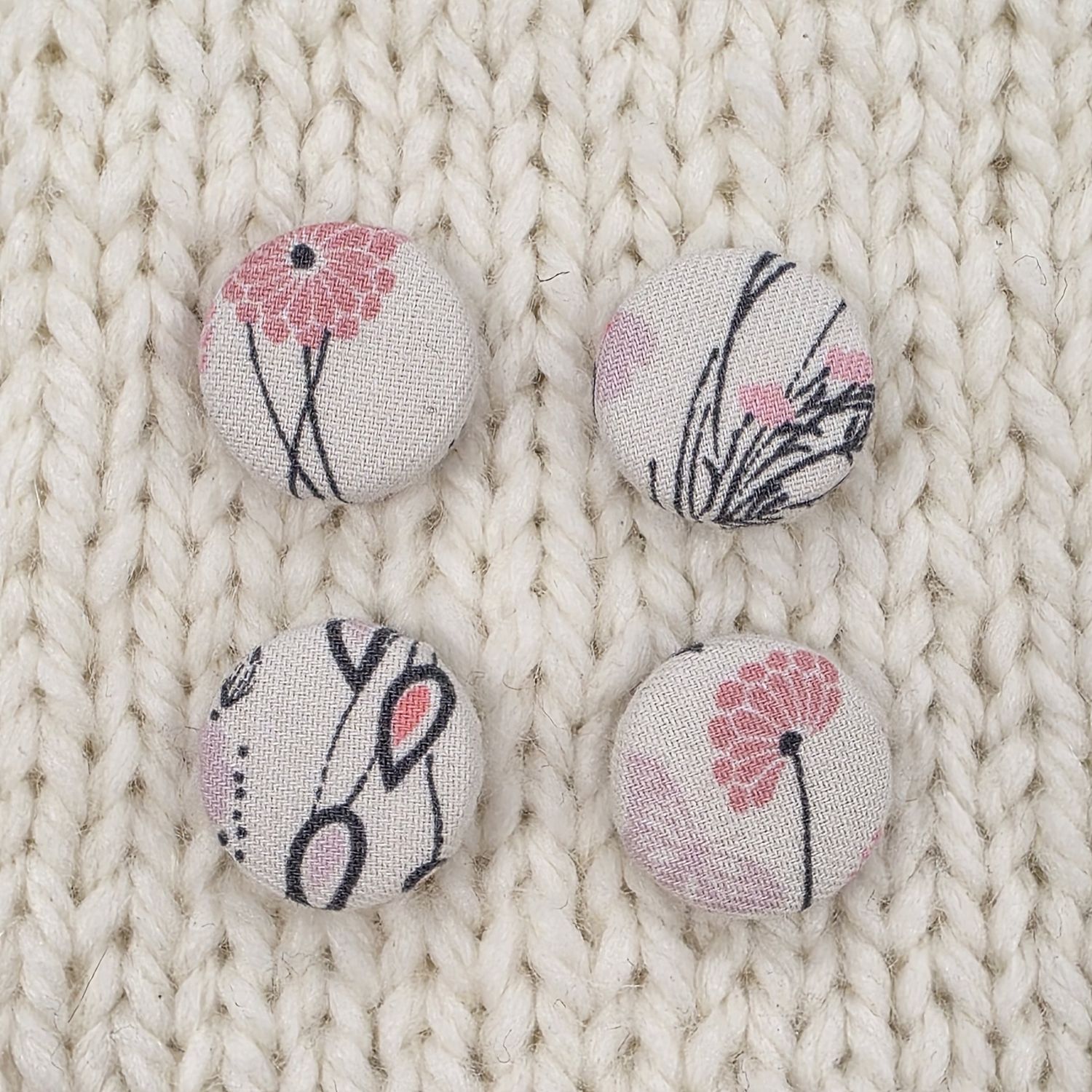4 white fabric covered buttons with grey and pink floral print, on a cream knitted background.