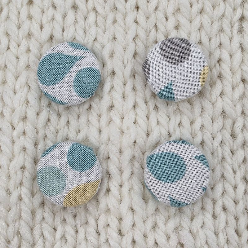 4 white fabric covered buttons with teal, yellow and grey raindrop print, on a cream knitted background.