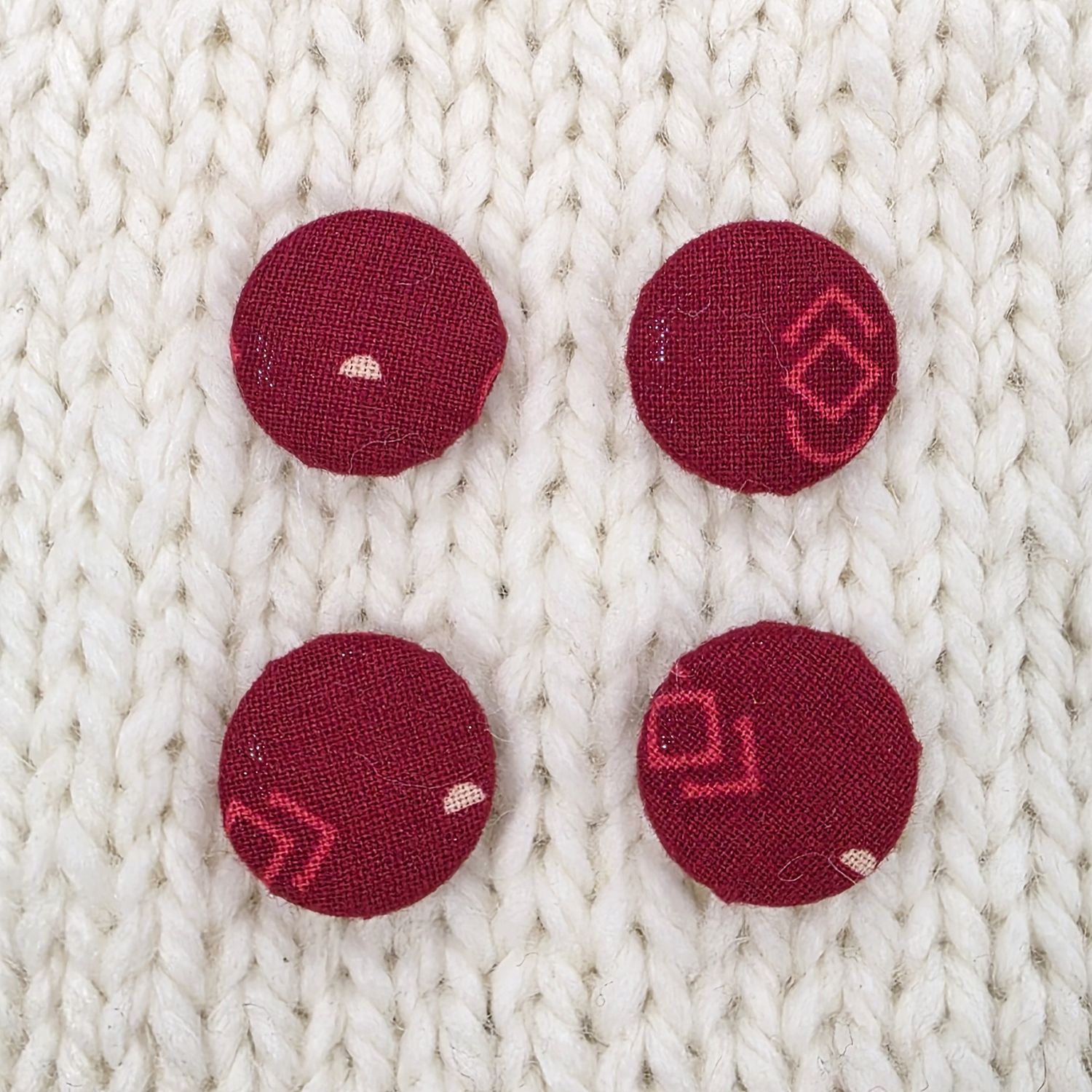 4 dark red fabric covered buttons with lighter red geometric print, on a cream knitted background.