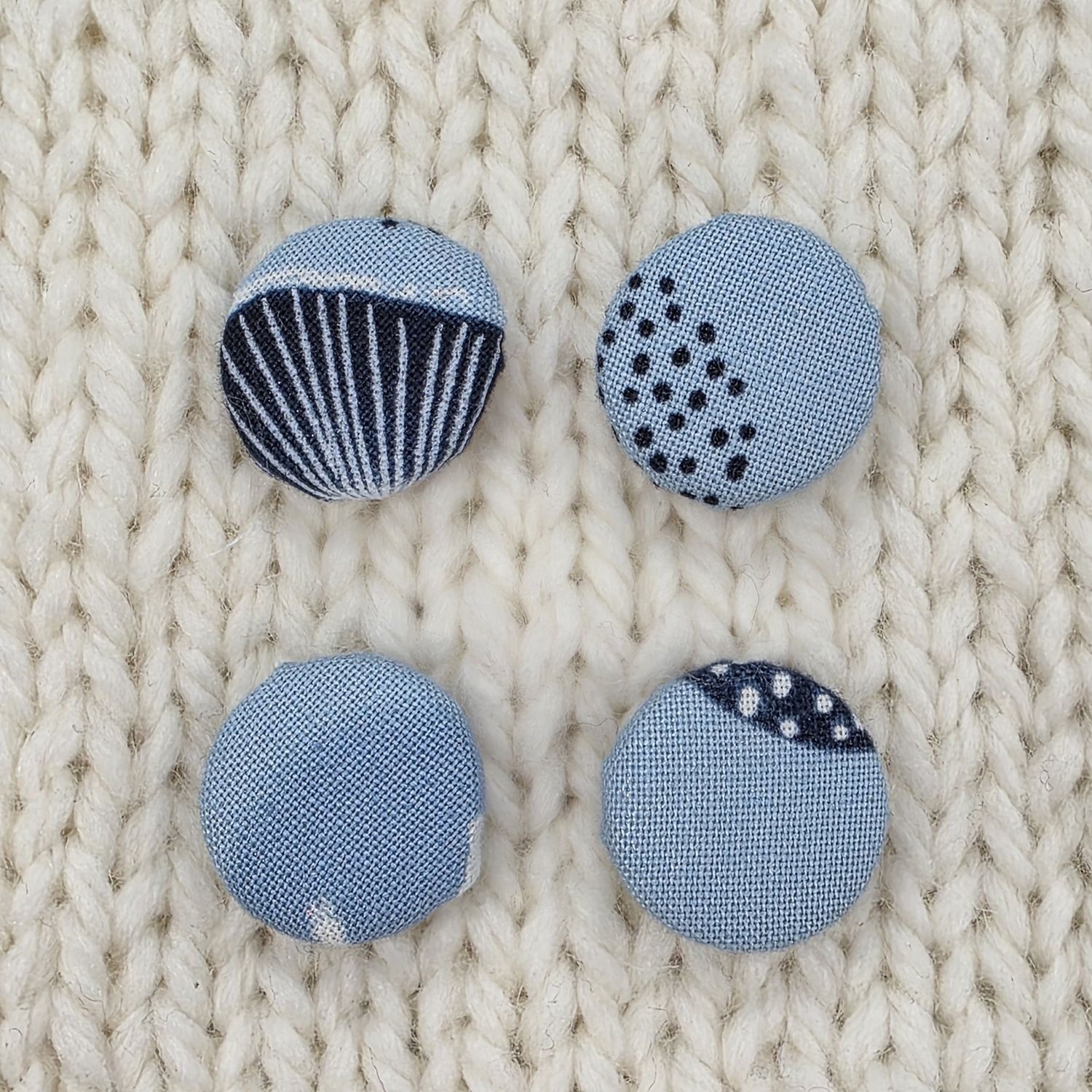 4 light blue fabric covered buttons with navy asbtract print, on a cream knitted background.