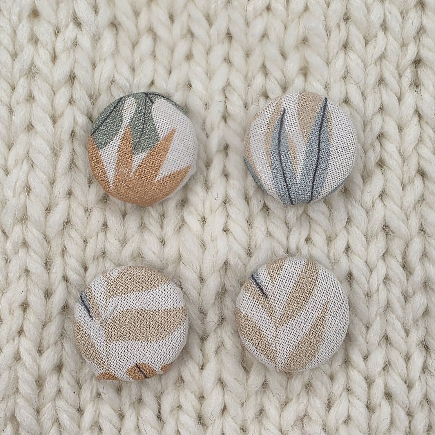 4 white fabric covered buttons with pale brown, blue and sage green leaf print, on a cream knitted background.