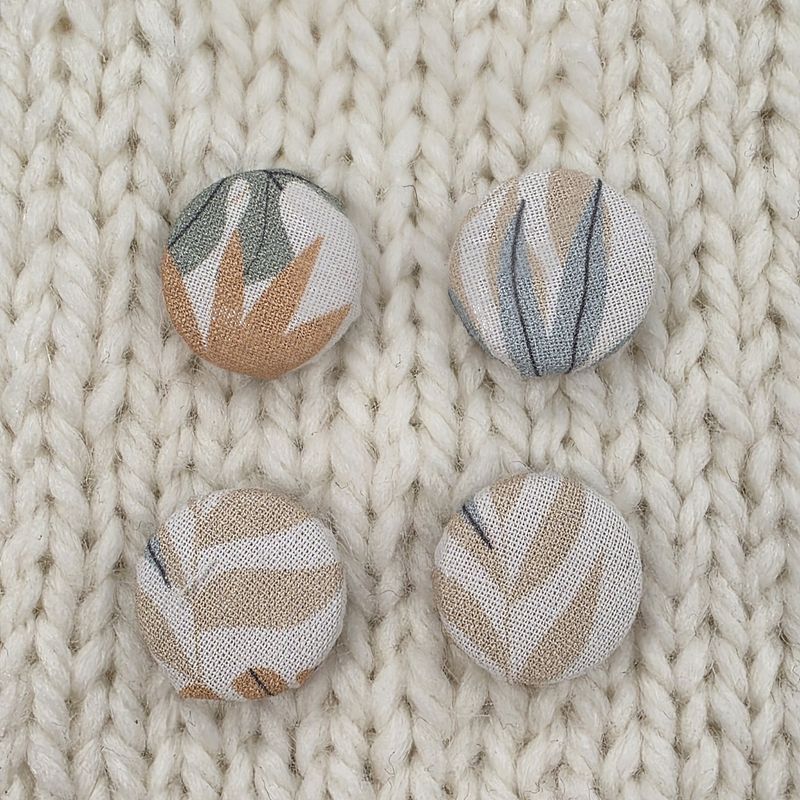 4 white fabric covered buttons with pale brown, blue and sage green leaf print, on a cream knitted background.