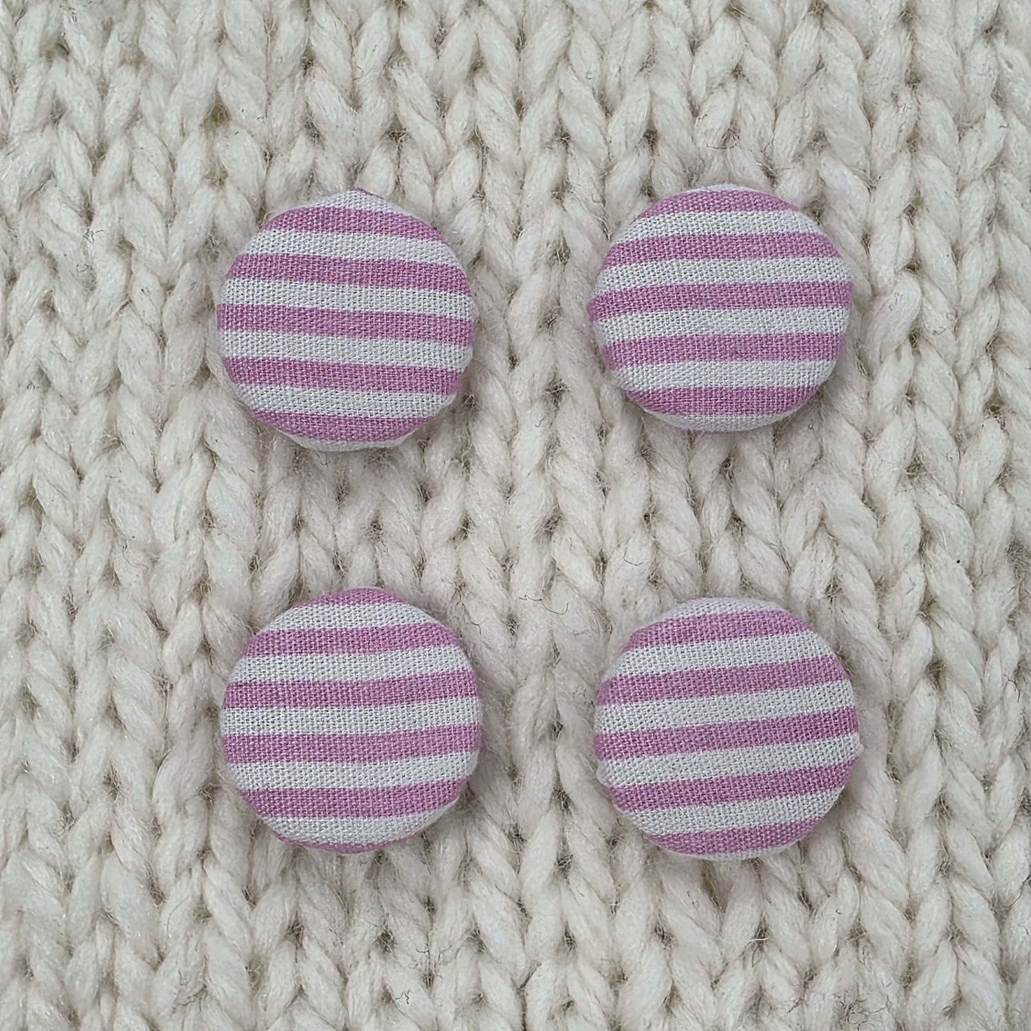 4 white fabric covered buttons with pink stripes, on a cream knitted background.