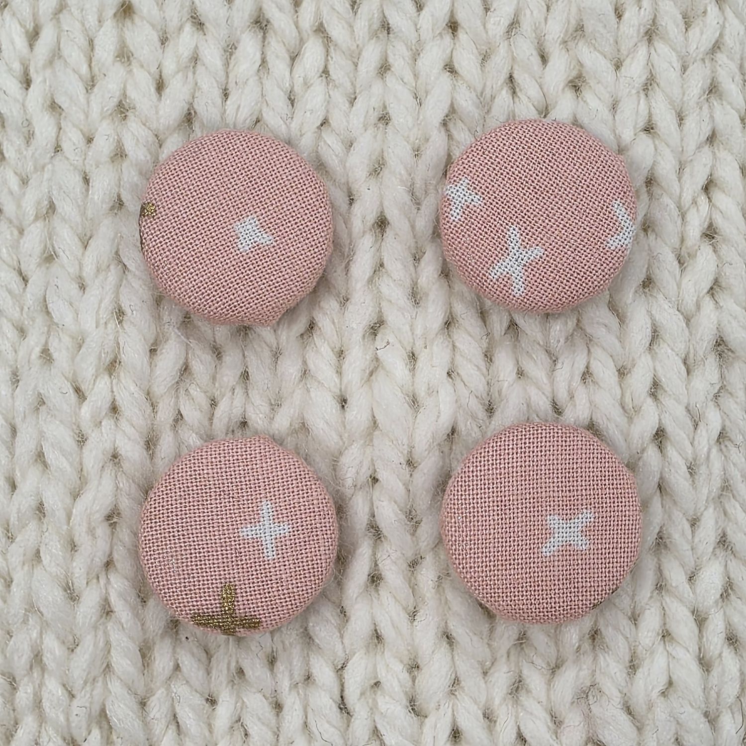 4 light pink fabric covered buttons with white and gold crosses print, on a cream knitted background.