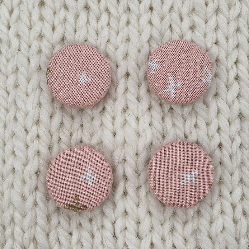 4 light pink fabric covered buttons with white and gold crosses print, on a cream knitted background.