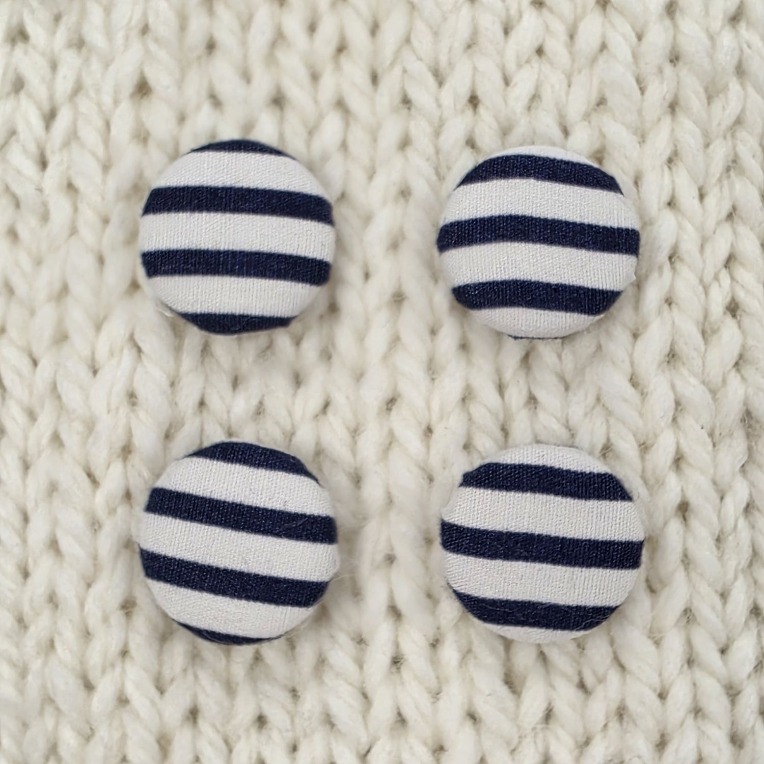4 white fabric covered buttons with navy stripes, on a cream knitted background.