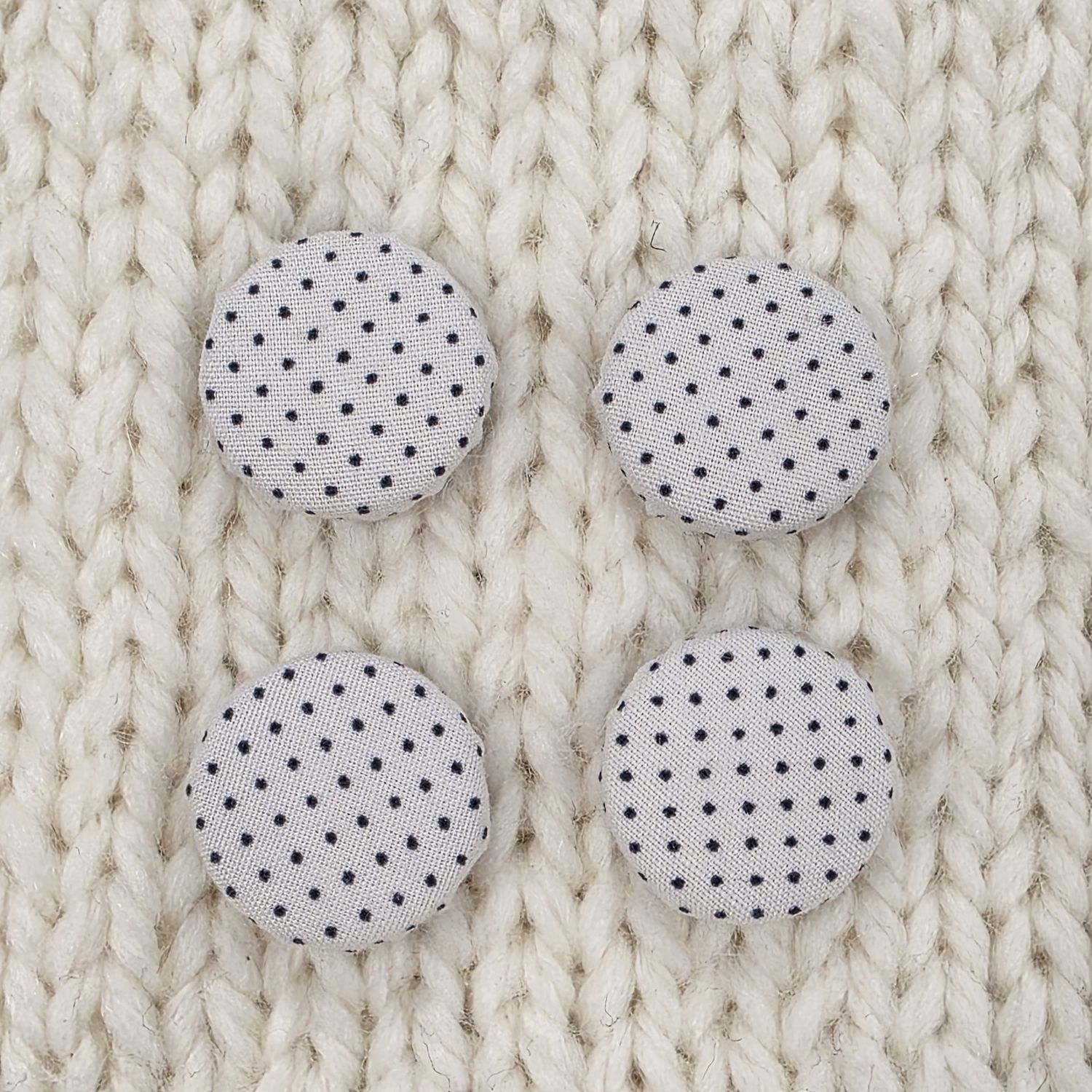 4 white fabric covered buttons with small black polka dot print, on a cream knitted background.
