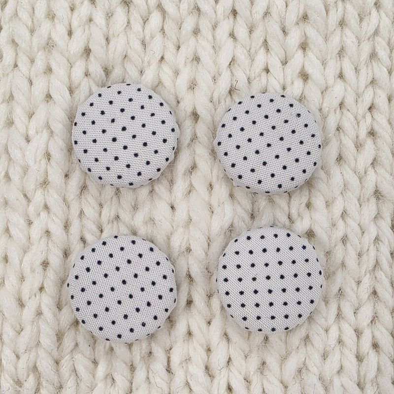 4 white fabric covered buttons with small black polka dot print, on a cream knitted background.