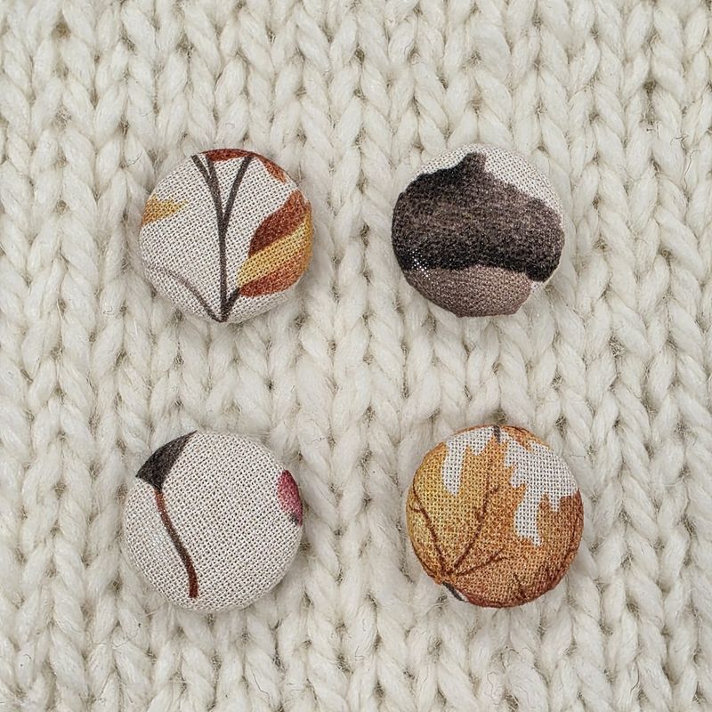 4 white fabric covered buttons with autumnal print such as leaves and acorns, on a cream knitted background.