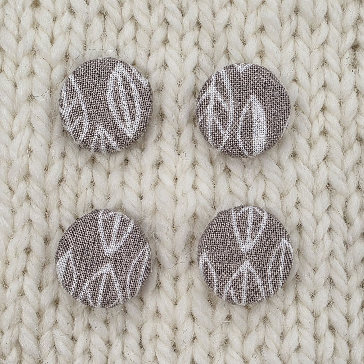 4 grey fabric covered buttons with white leaf print, on a cream knitted background.