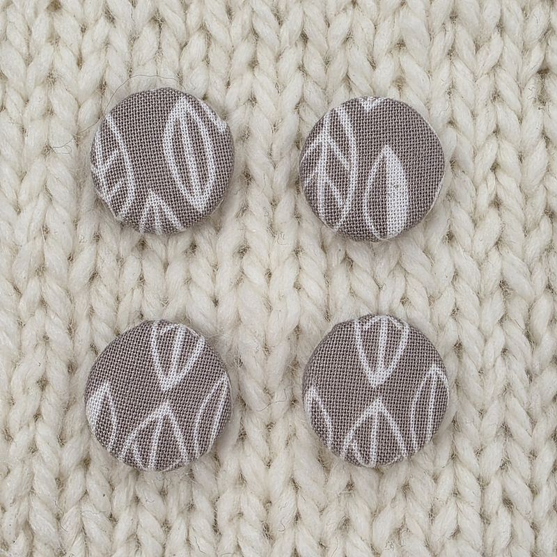 4 grey fabric covered buttons with white leaf print, on a cream knitted background.