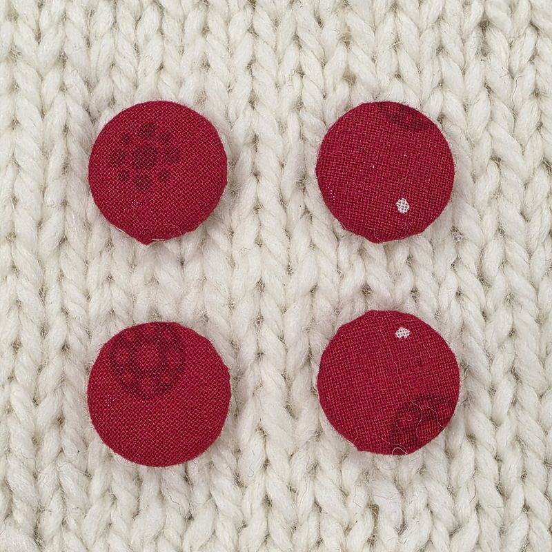 4 red fabric covered buttons with a dark red circular print, on a cream knitted background.