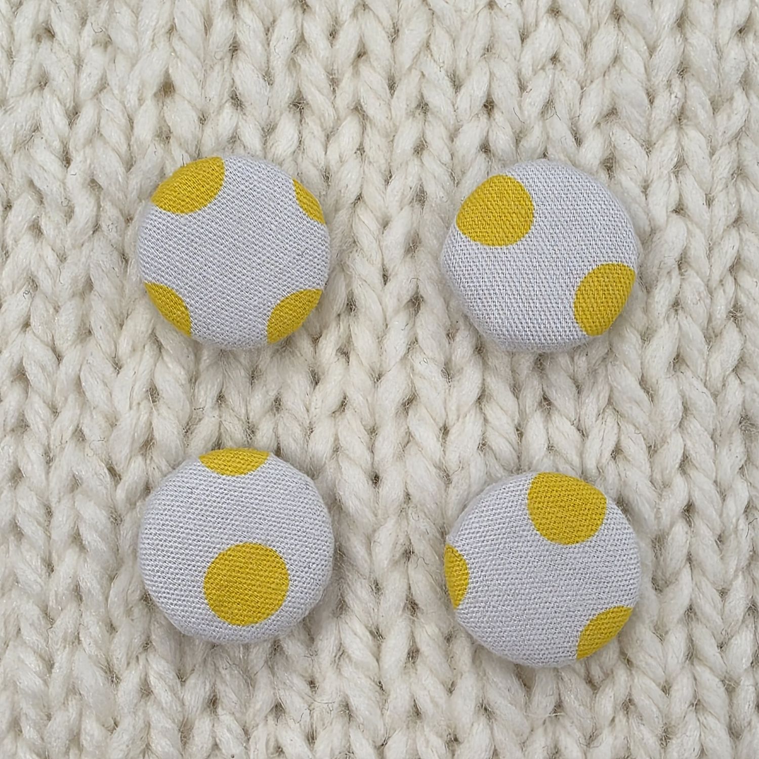 4 white fabric covered buttons with yellow dotty print, on a cream knitted background.