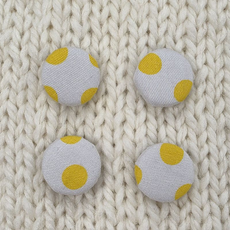 4 white fabric covered buttons with yellow dotty print, on a cream knitted background.