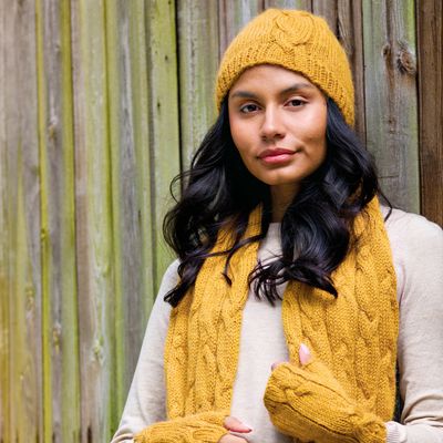 A woman is wearing a mustard yellow cable knit hat, scarf and wrist warmers.