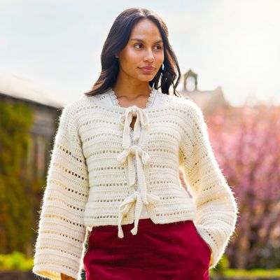 A woman is wearing a cream crocheted cardigan with ties on the front.