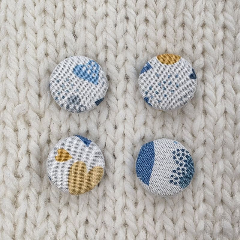 4 white fabric covered buttons with blue and yellow heart print, on a cream knitted background.
