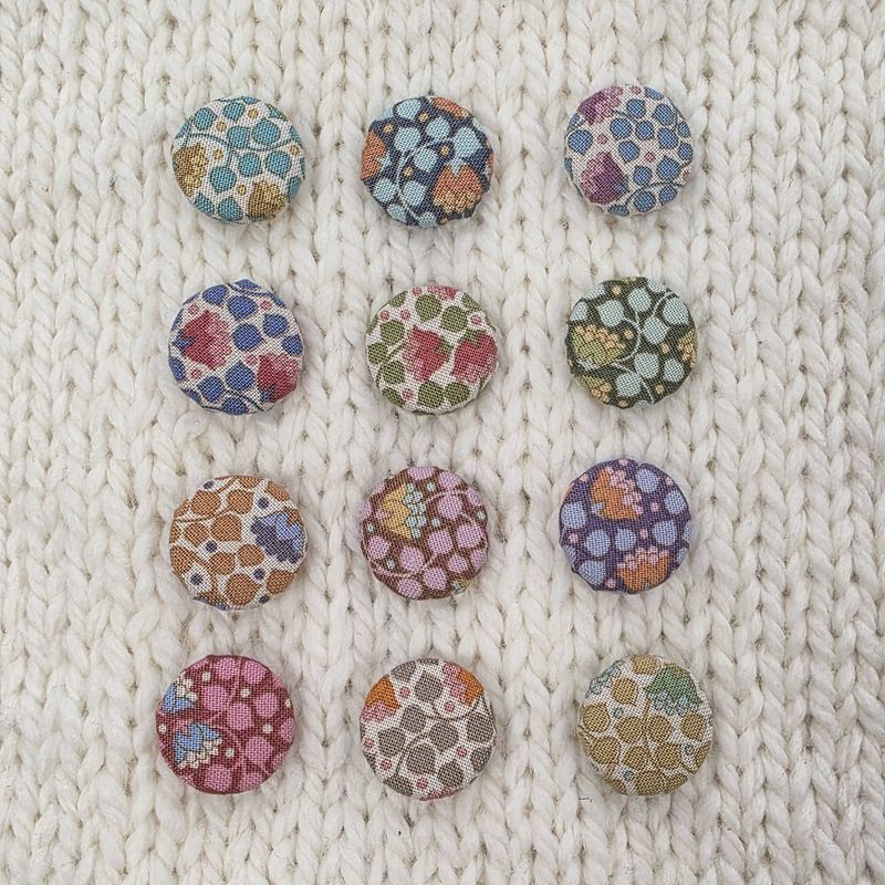 12 fabric covered buttons with various colours of floral print, on a cream knitted background.