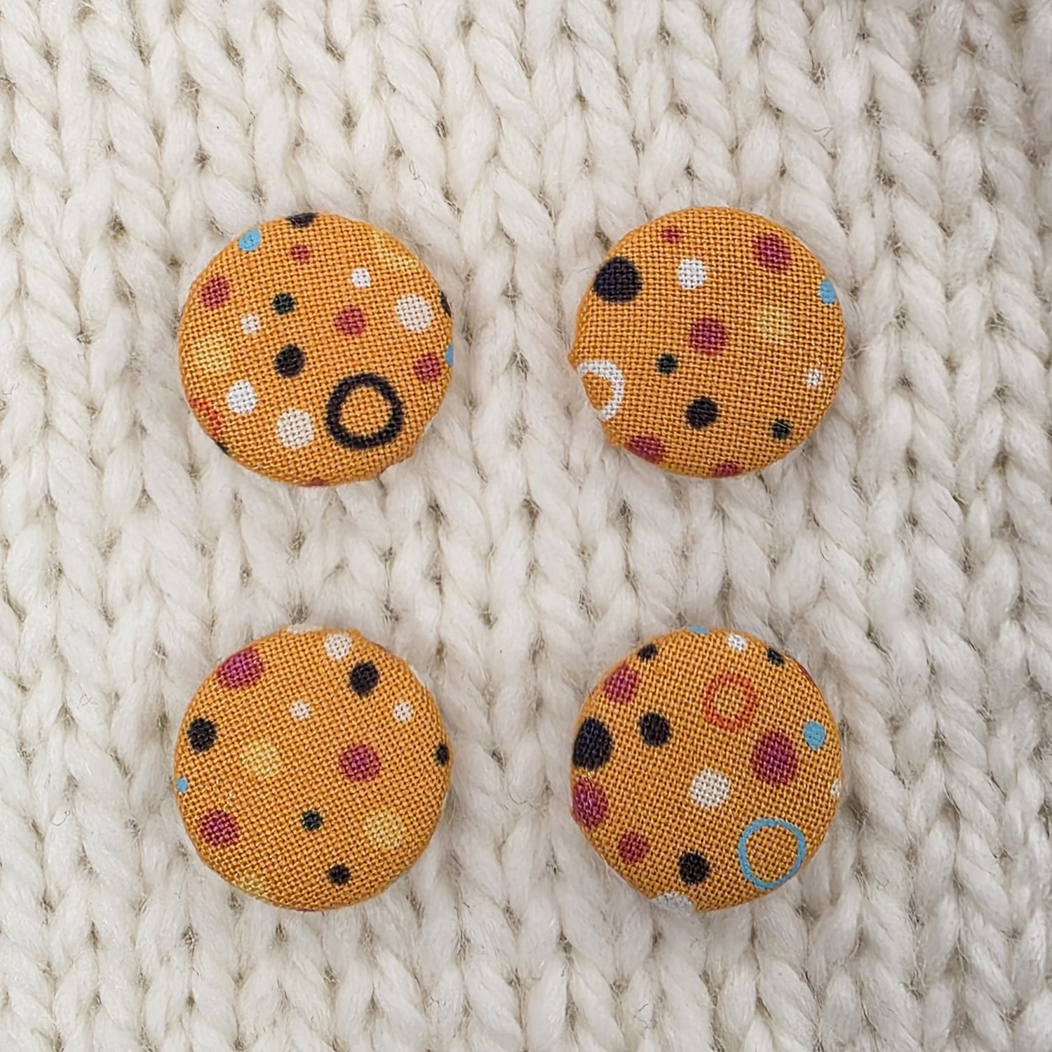 4 orange fabric covered buttons with multicoloured spots on a cream knitted background.
