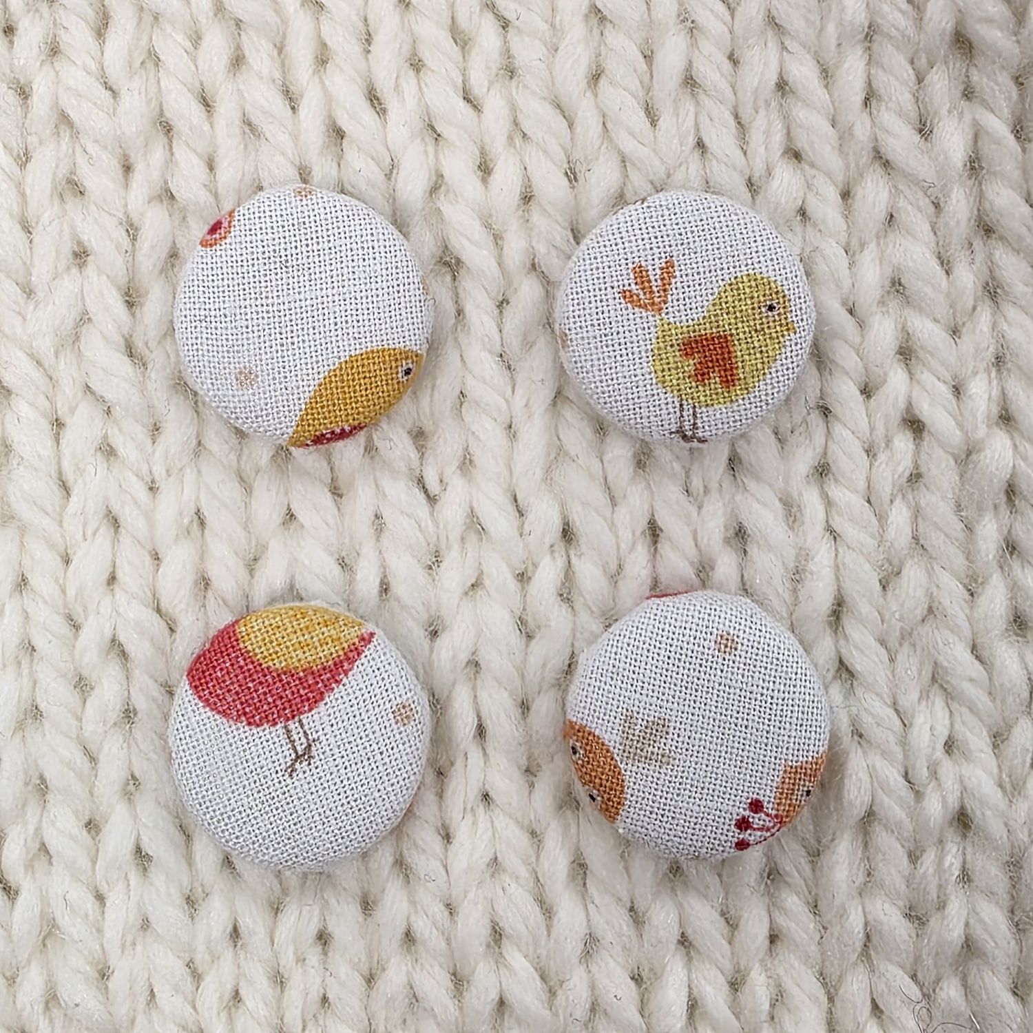 4 white fabric covered buttons with red and yellow cartoon bird print, on a cream knitted background.