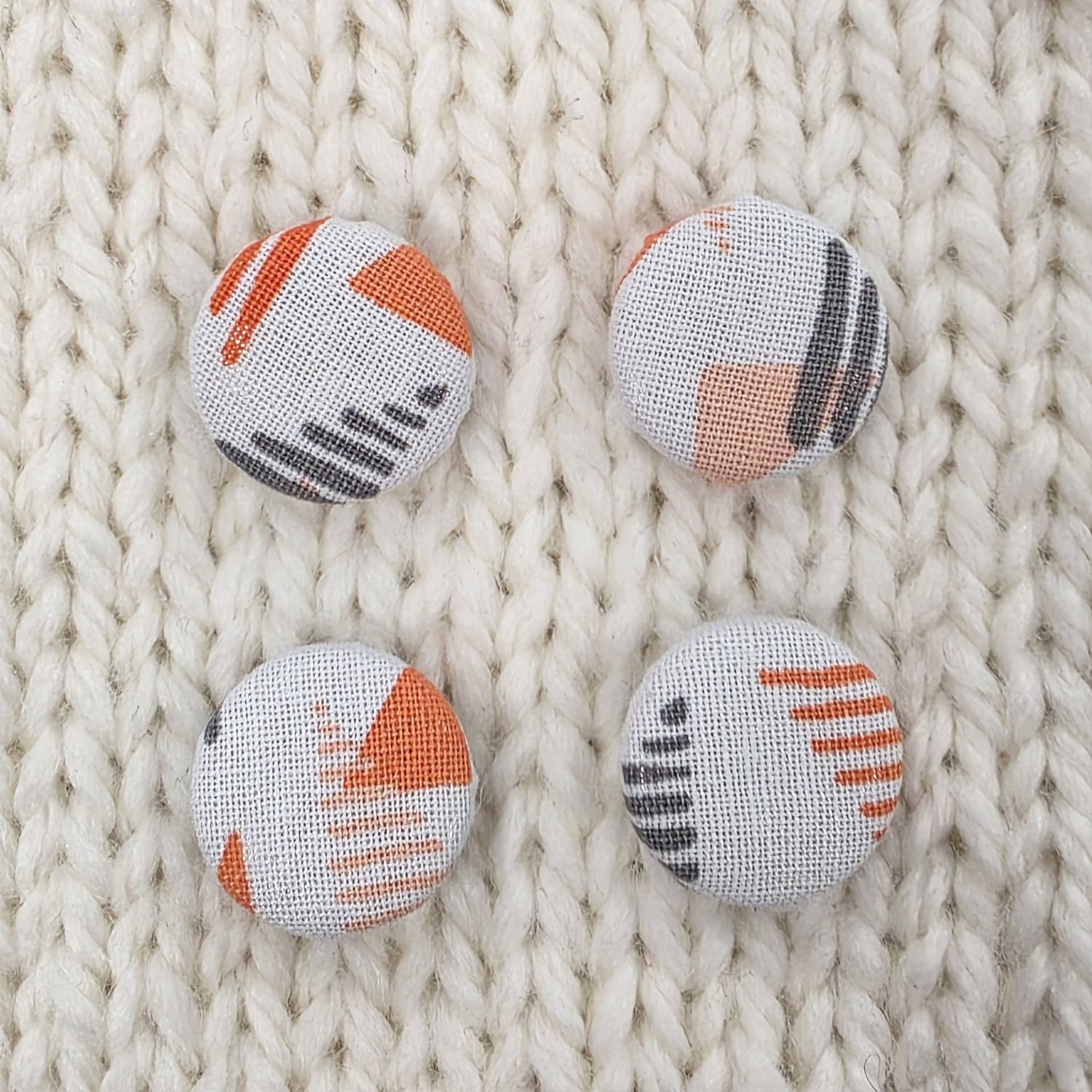 4 white fabric covered buttons with orange and grey geometric triangle print, on a cream knitted background.
