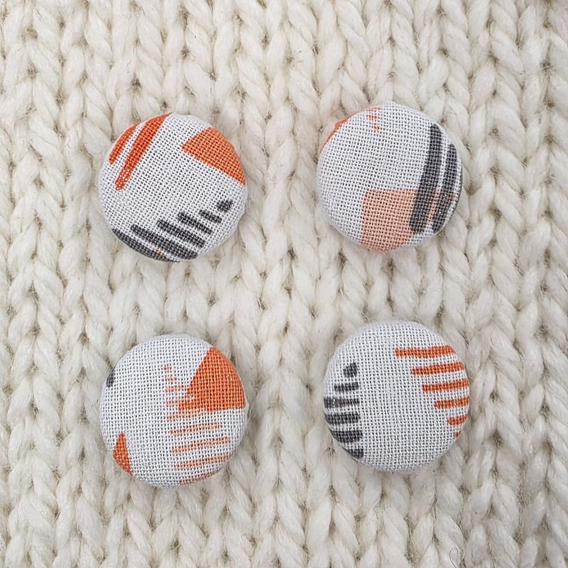 4 white fabric covered buttons with orange and grey geometric triangle print, on a cream knitted background.