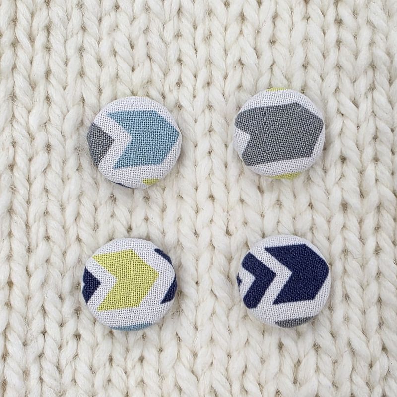 4 white fabric covered buttons, with grey, green and blue chevron print on a cream knitted background.