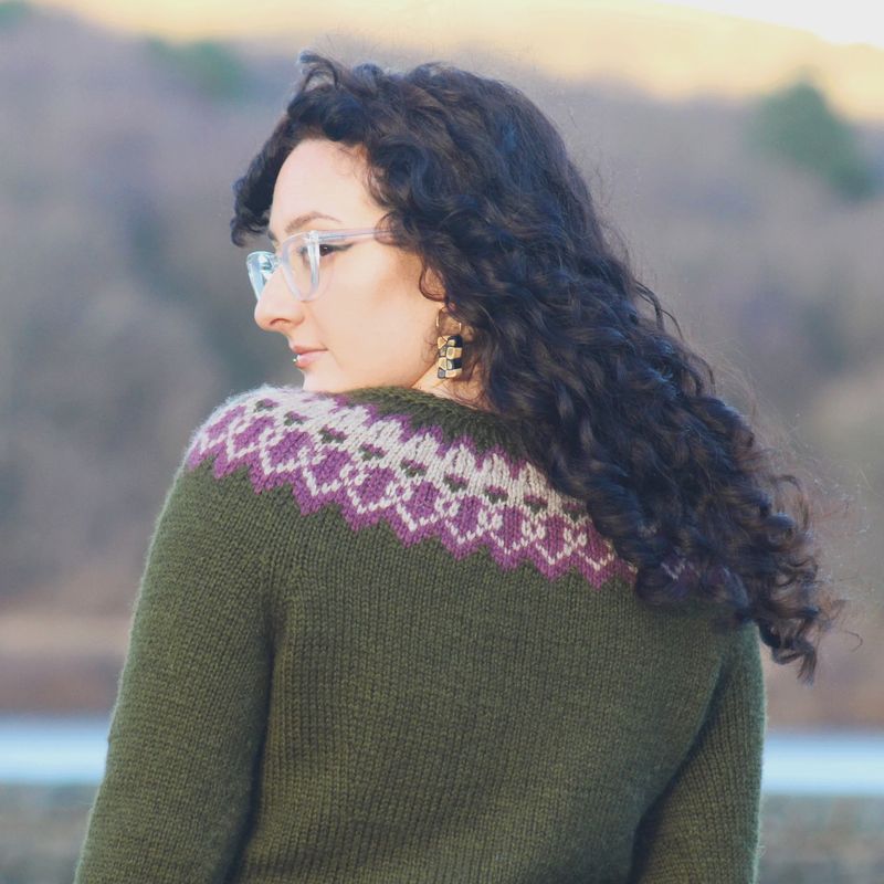 A woman is wearing a dark green knitted sweater with cream and purple colourwork around the yoke.