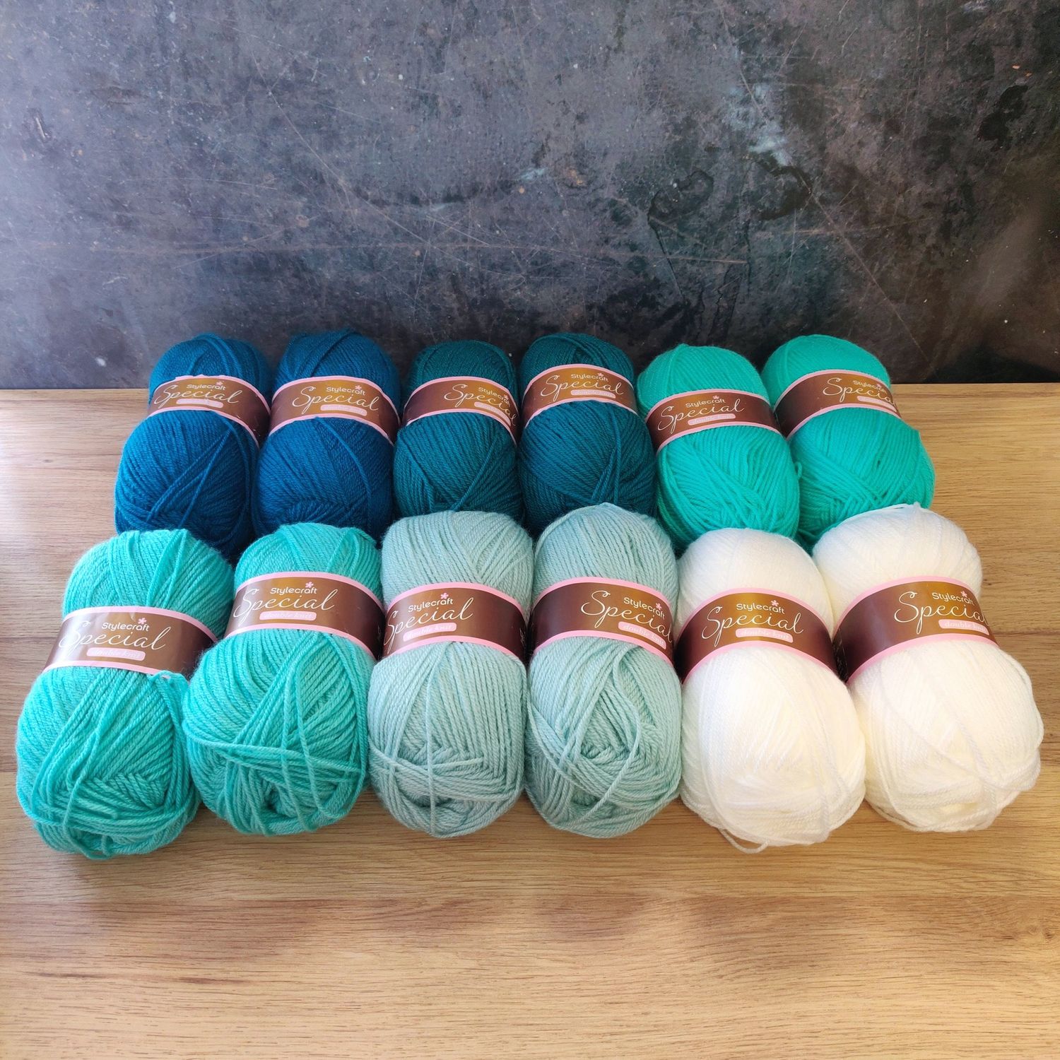 12 balls of yarn in shades of teal, aqua and white laid out on a wooden surface.