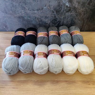 12 balls of yarn in shades of black, grey, silver and white laid out on a wooden surface.