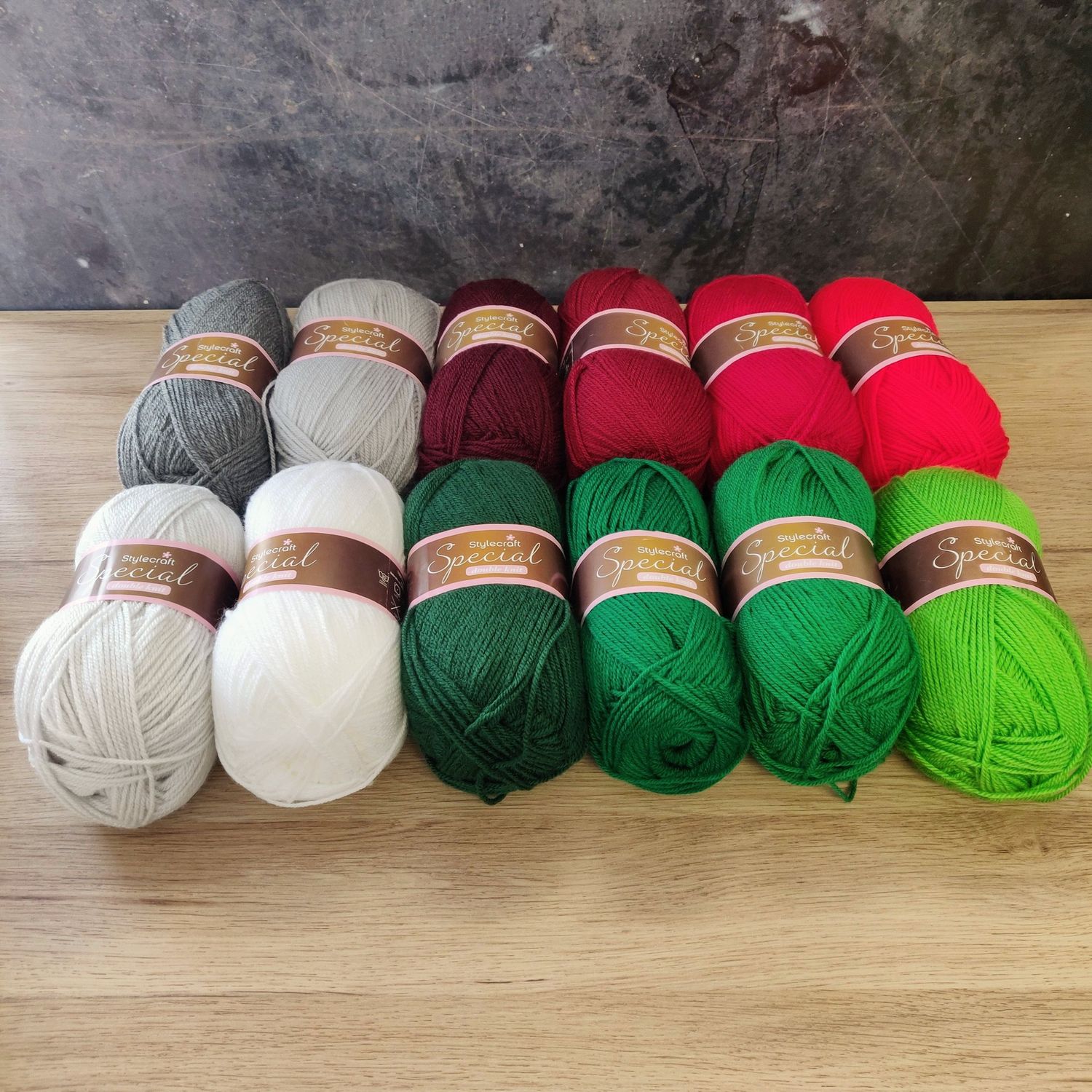 12 balls of yarn in shades of grey, white, red and green laid out on a wooden surface.