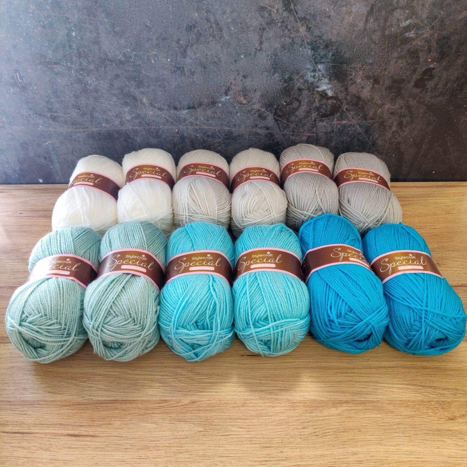 12 balls of yarn in shades of white, silver and blue laid out on a wooden surface.