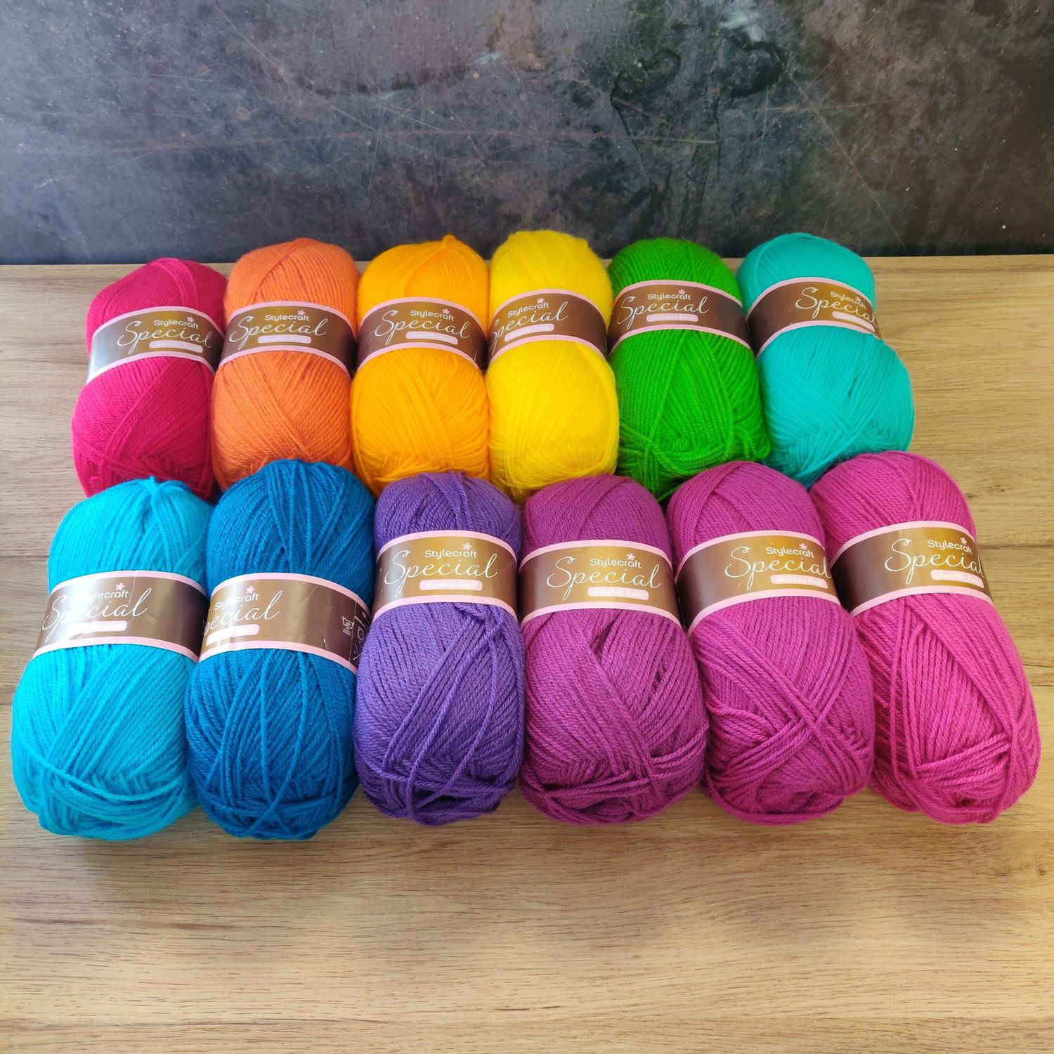 12 balls of yarn in shades of red, orange, yellow, green, teal, blue, purple and pink laid out on a wooden surface.